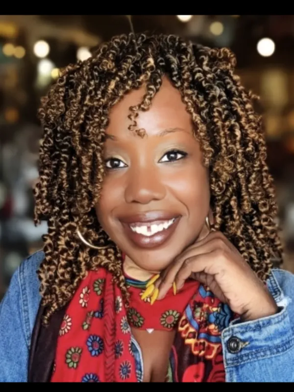 African American woman with brown curly hair, wearing gold hoops, a jean jacket, and multicolor red scarf with flowers 